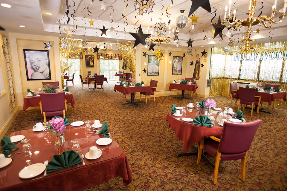 A decorated dining room with maroon tablecloths and purple chairs. Tables are set with white plates, cups, glasses, and green folded napkins. The ceiling is adorned with hanging black and gold star decorations. The walls feature framed pictures of classic Hollywood stars, and a gold tinsel curtain with the word 'HOLLYWOOD' is visible in the background.