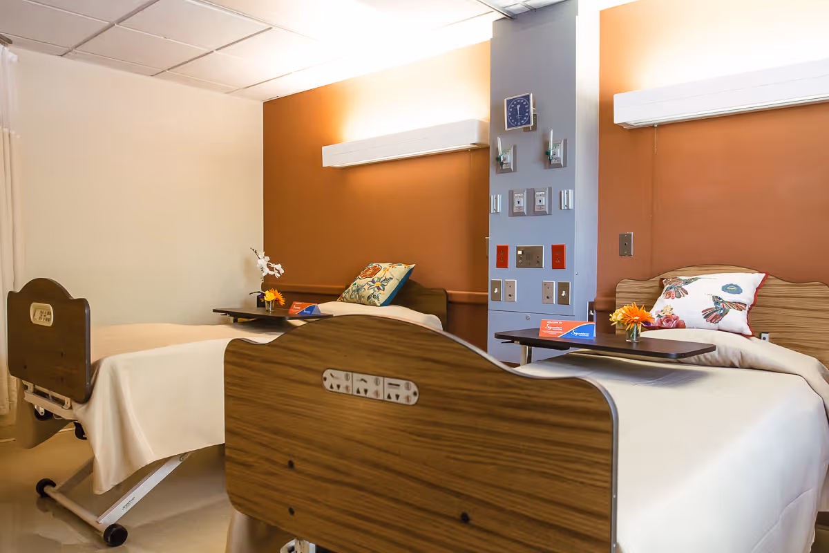 A room with two hospital-style beds, each with a floral pillow and a small table holding a flower vase. The walls are painted in a warm orange and beige color, and there are medical equipment panels mounted on a central column between the beds.