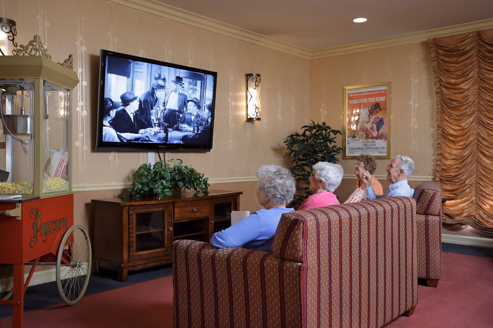Four elderly people sitting on patterned couches in a cozy living room watching a black and white movie on a wall-mounted TV. There is a vintage popcorn machine on the left, a wooden cabinet with a plant, a movie poster on the wall, and decorative curtains on the right.