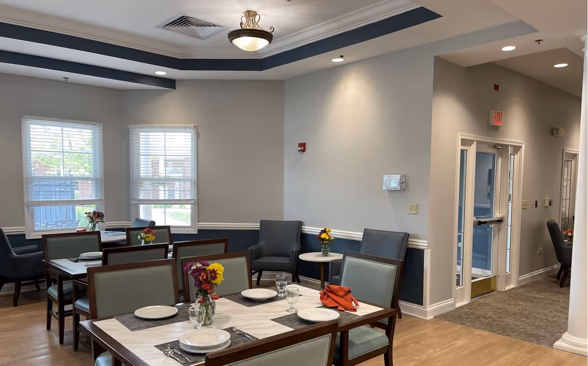 A dining room in a senior living facility with several tables set with plates, glasses, and napkins. Each table has a small vase with colorful flowers. The room has light gray walls with a darker blue wainscoting, two windows with white blinds, and a ceiling light fixture. There are upholstered chairs around the tables and a hallway with a glass door leading to another area.