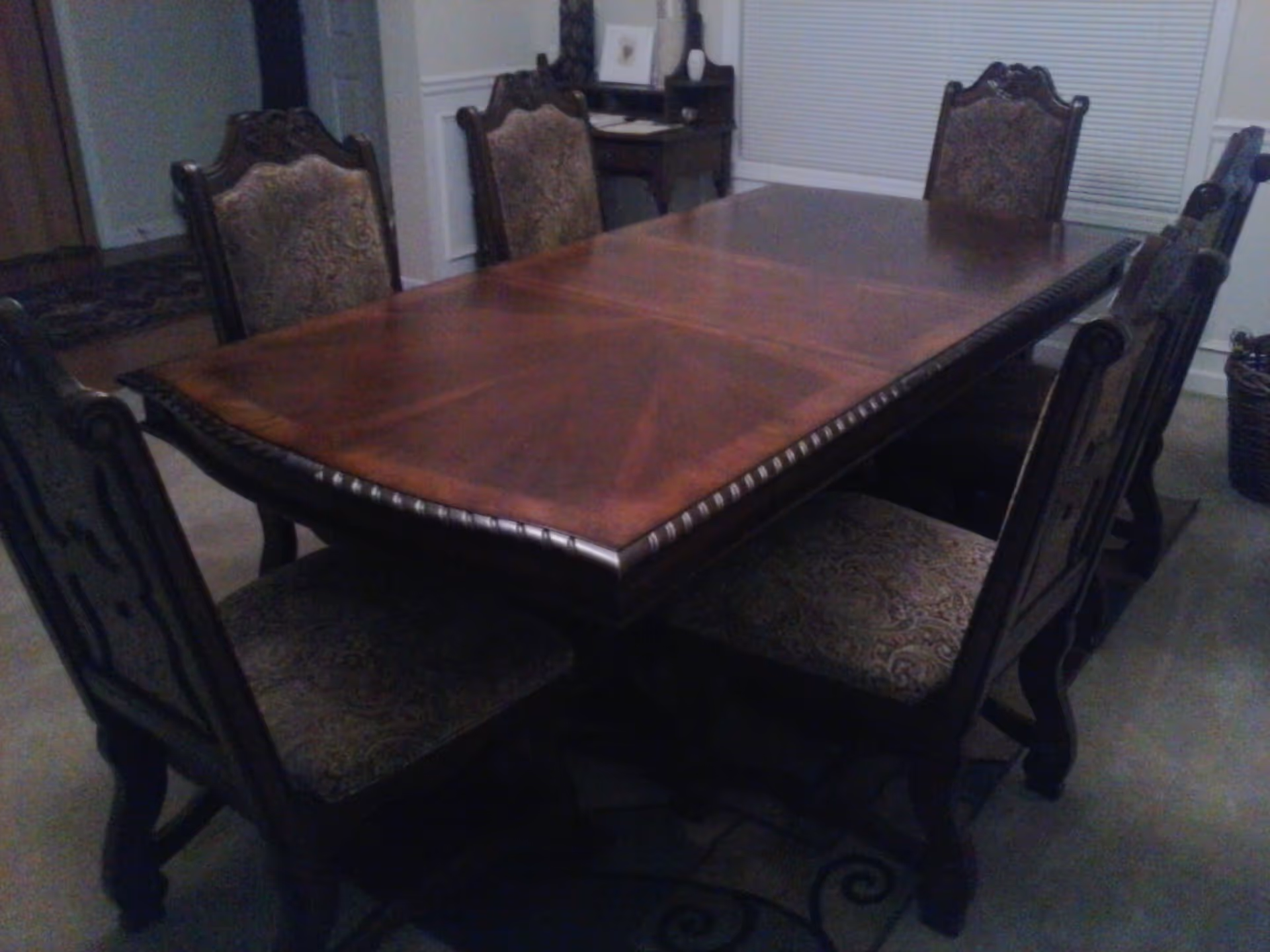 A wooden dining table with six upholstered chairs arranged around it in a dining room. The room has a carpeted floor, a window with closed blinds, and a small desk with items on it in the background.
