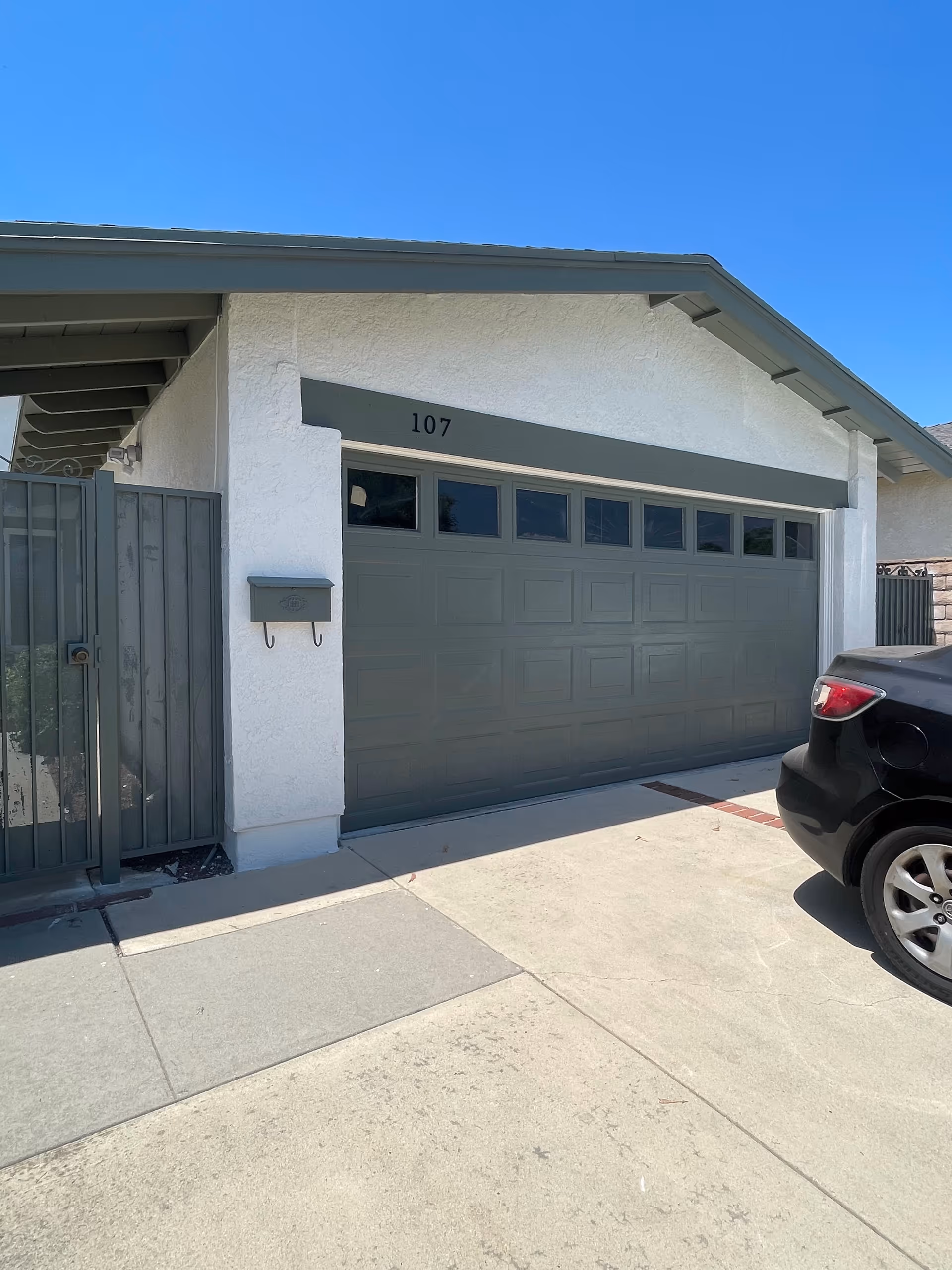 Front exterior of a house showing a gray garage door numbered 107 with a gate, mailbox, and a parked car.