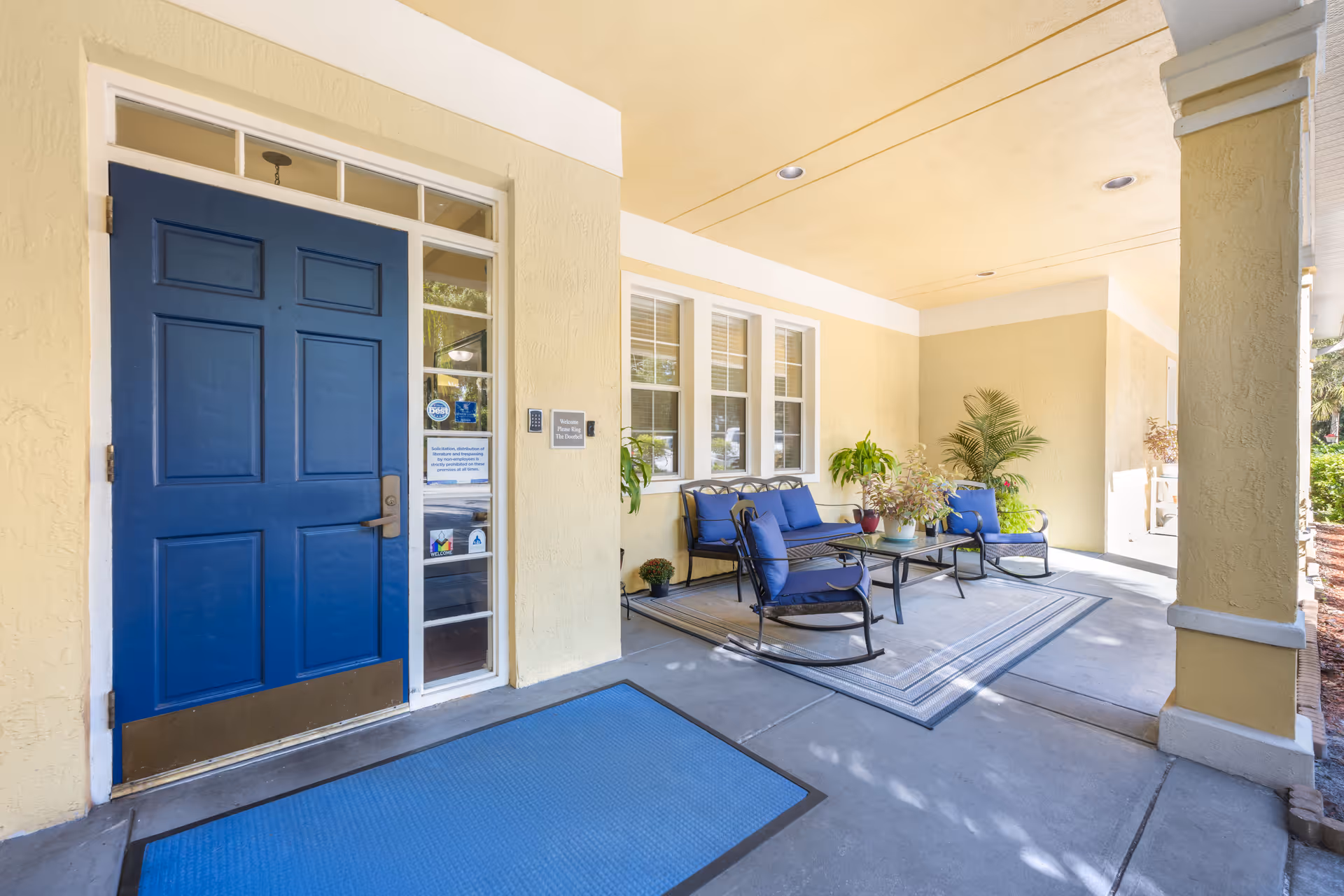 Covered outdoor seating area with a blue door on the left, a blue mat on the floor, and a seating arrangement consisting of a bench and two chairs with blue cushions around a glass-top table. Several potted plants are placed near the seating area, and the walls are painted yellow with white trim.