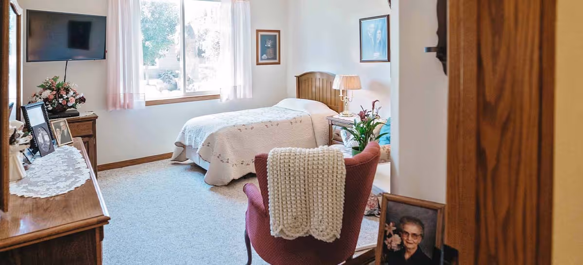 A cozy bedroom with a single bed covered in a white quilt, a wooden headboard, and a bedside table with a lamp and flowers. There is a red armchair with a knitted blanket draped over it, a wooden dresser with framed photos and a floral arrangement, and a wall-mounted TV. The room has a large window with sheer curtains letting in natural light and framed pictures on the walls.