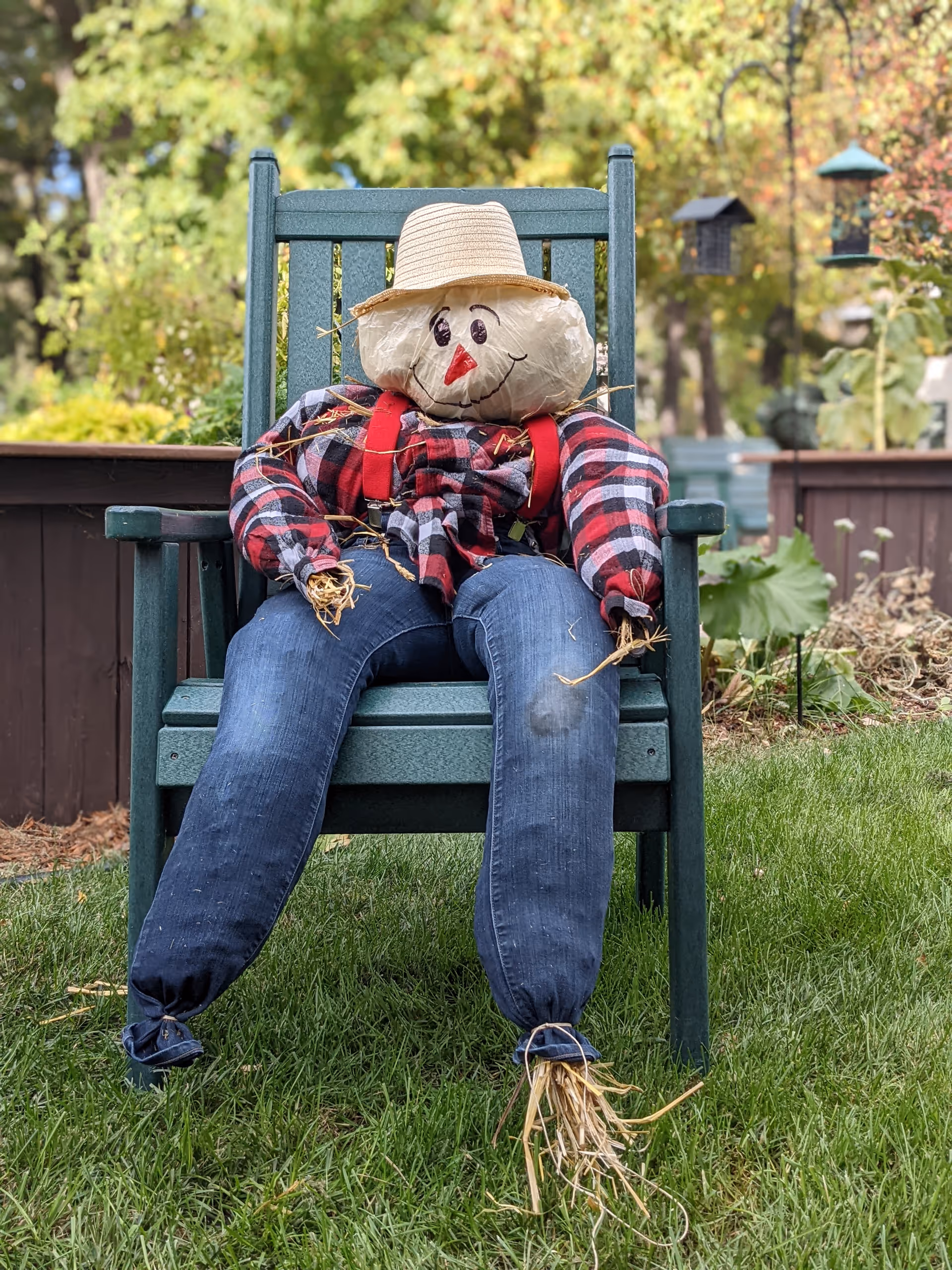 A scarecrow wearing a straw hat, plaid shirt and jeans sits slumped on a green wooden chair in a grassy yard.