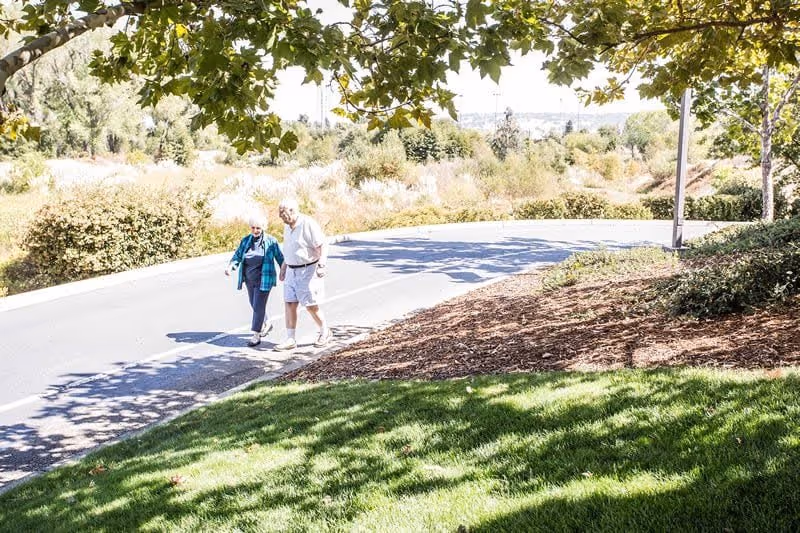 Two elderly people walking together on a paved path surrounded by greenery and trees on a sunny day at Creekside Oaks Retirement Community.