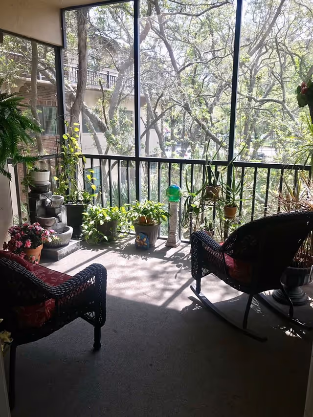 Sunlit screened balcony with wicker rocking chairs, numerous potted plants, and a view of trees outside.