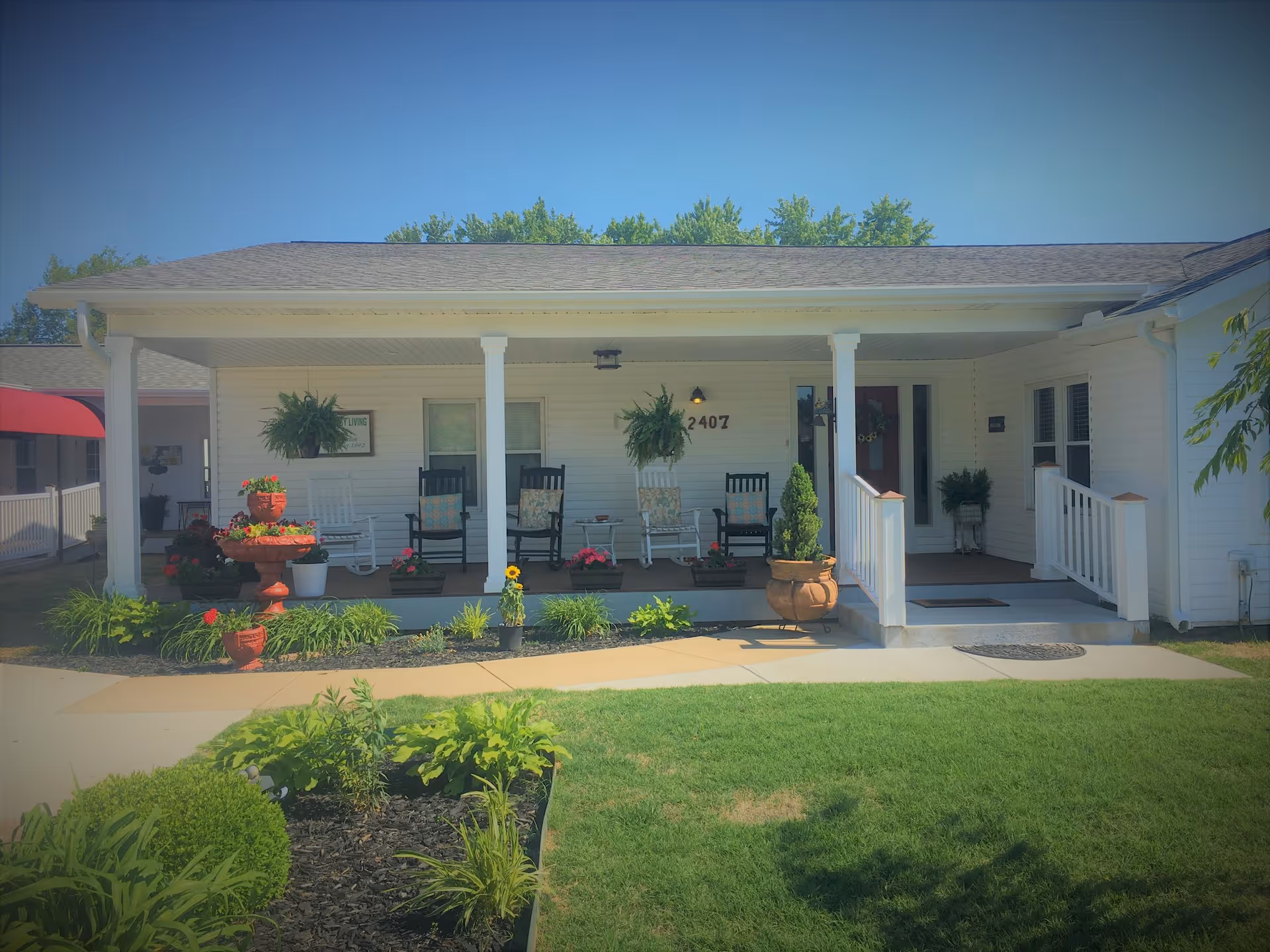 Front porch of a single-story white building with rocking chairs, potted plants, and the address "2407" visible, facing a green lawn.