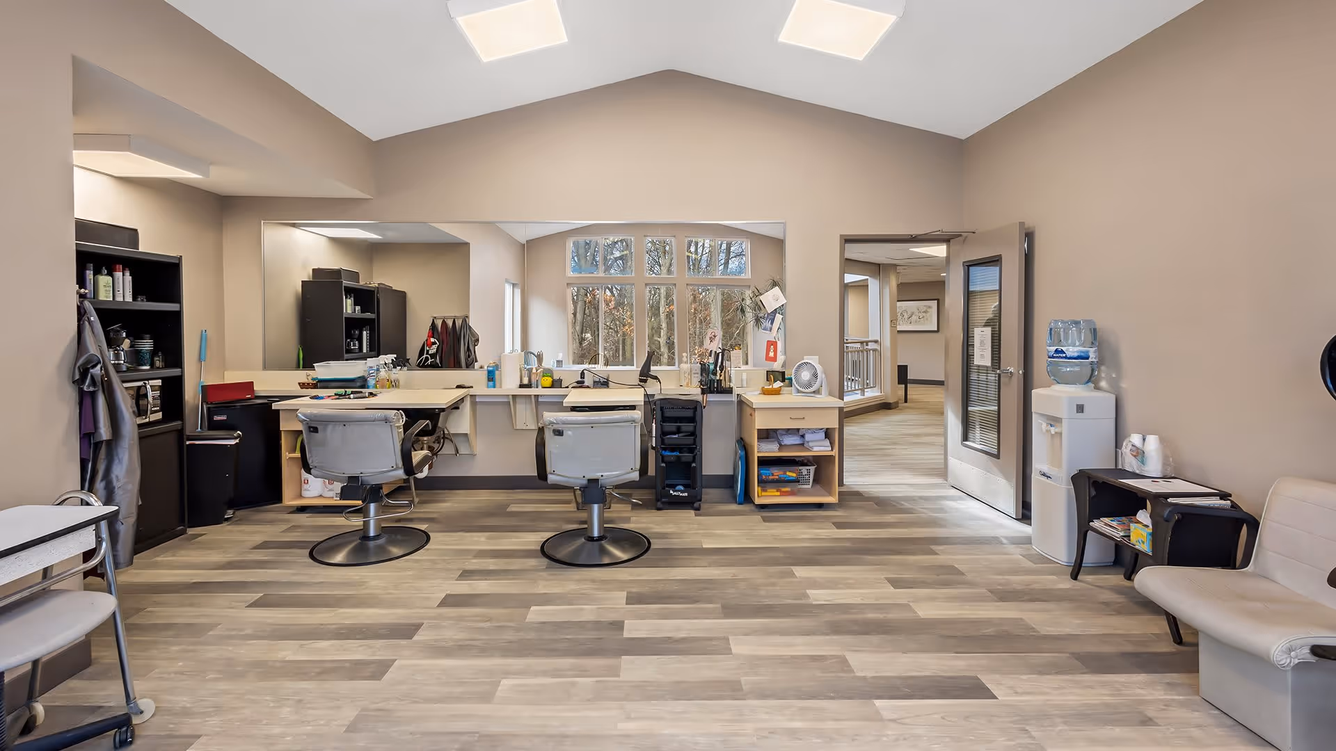 Interior view of a salon area in a senior living facility with two salon chairs in front of a large mirror, shelves with hair care products, a water cooler, and seating area with beige chairs. The room has wood-look flooring and beige walls with a window showing trees outside.