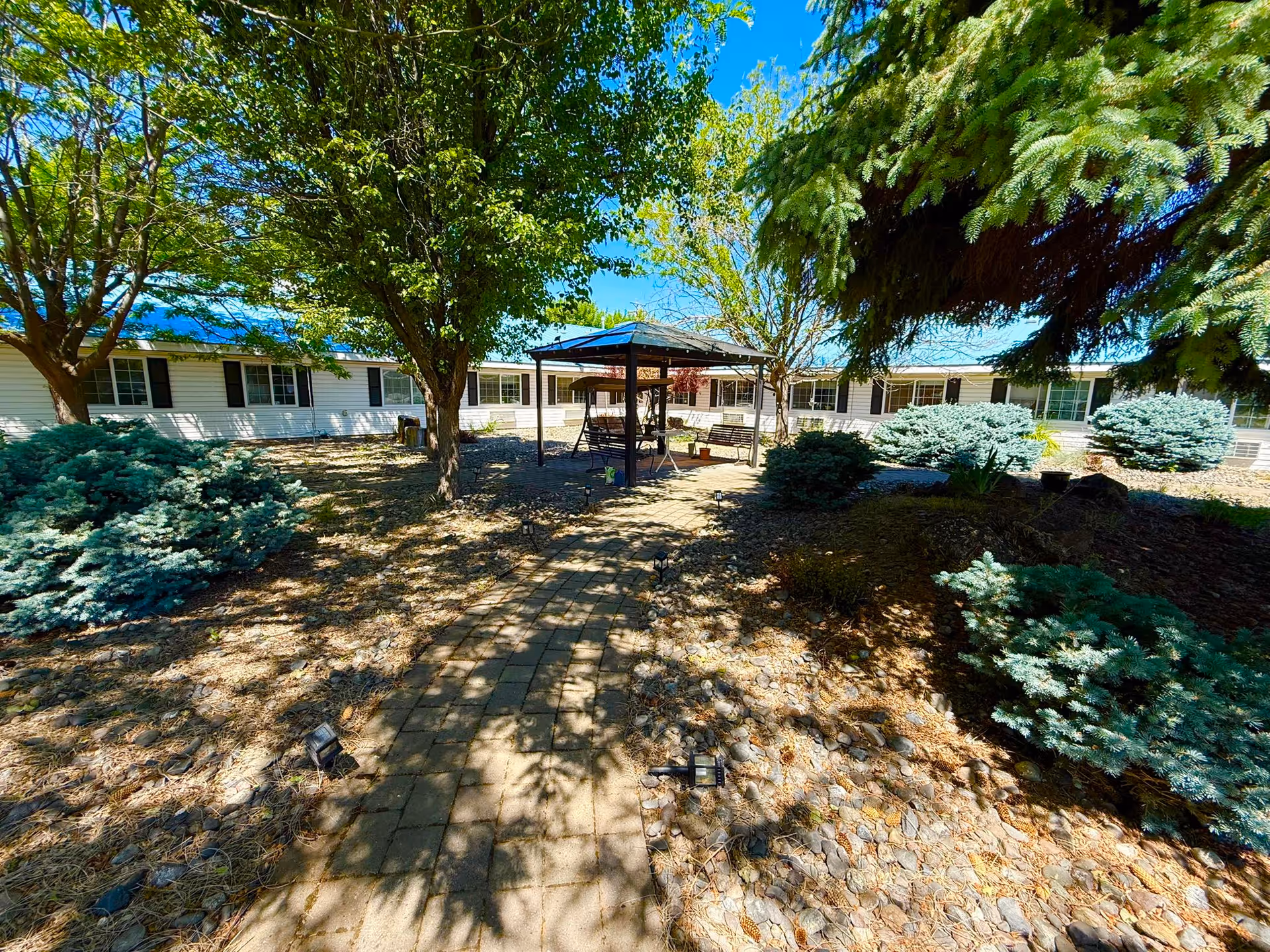 Outdoor garden area at Haven House Retirement Center with a paved walkway leading to a gazebo with seating. The area is surrounded by trees, shrubs, and bushes under a clear blue sky.