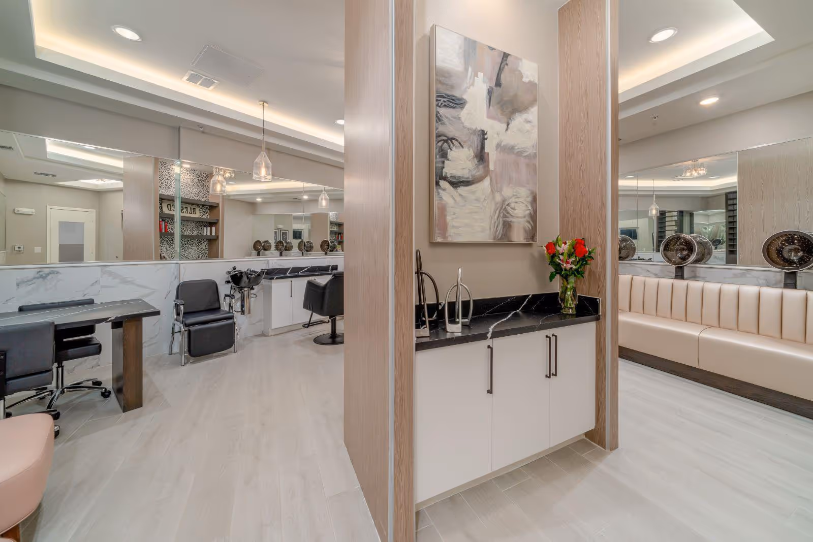 Interior view of a modern salon area in a senior living facility featuring multiple black salon chairs, hair washing stations, a manicure table with chairs, and a beige cushioned bench along the wall. The space has light wood flooring, large mirrors, pendant lighting, and a decorative abstract painting above a black countertop with a vase of flowers.