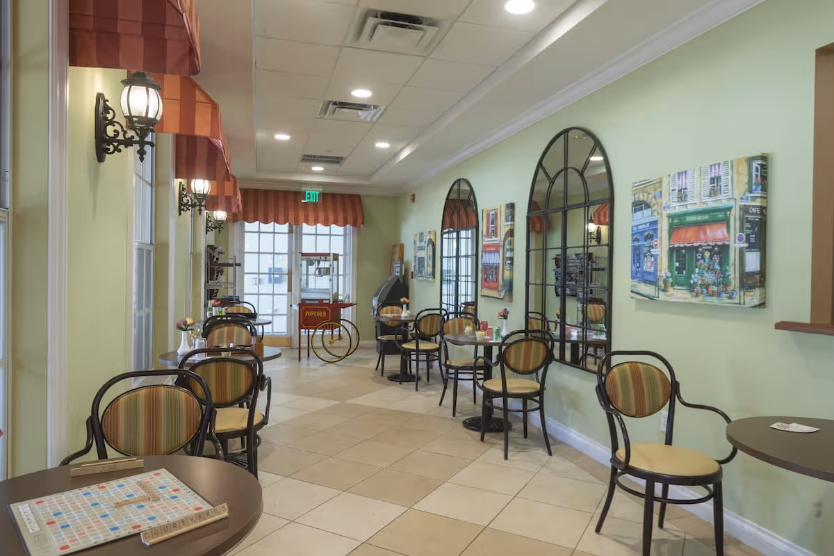 Bright communal dining/cafe area with small round tables and striped chairs along a pale green wall decorated with mirrors and paintings.