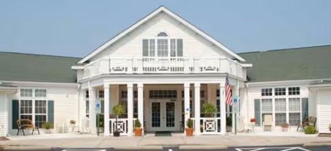 White, two-story senior living facility with a front entrance, columns, potted plants, and an American flag.
