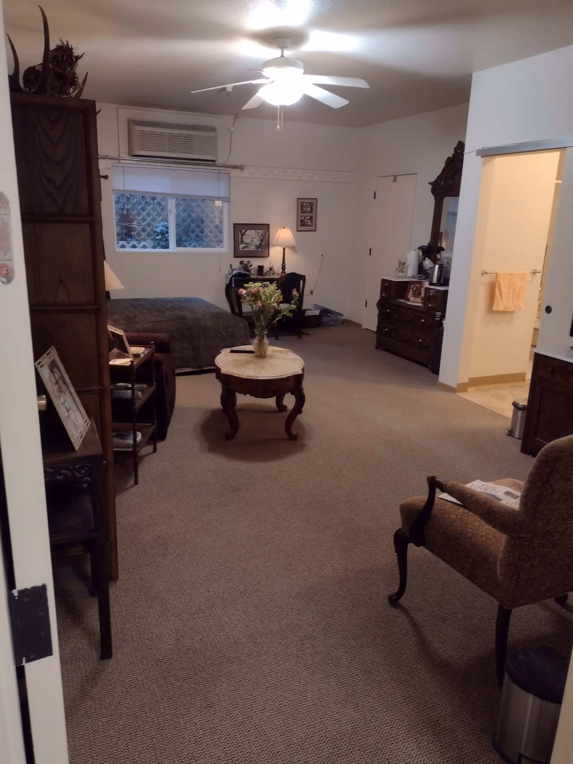 A cozy assisted living room with a bed near a window, a ceiling fan with light, a small round table with a vase of flowers, an armchair, a dresser with a mirror, and a doorway leading to a bathroom with a towel hanging.