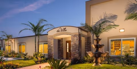 Exterior view of a modern facility entrance with stone archway, palms, and landscaped grounds.