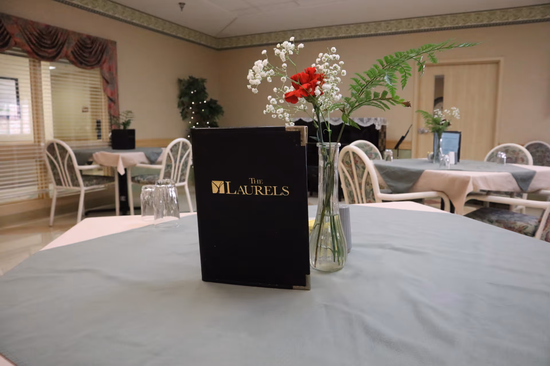 A dining room with tables covered in light-colored tablecloths and chairs arranged around them. On the closest table, there is a black menu with 'The Laurels' written in gold letters and a glass vase holding a red flower, white baby's breath, and green fern leaves. The room has beige walls, a window with blinds and a valance, and some plants in the background.