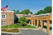 Exterior view of Calvert County Nursing Center showing a brick building with an American flag on a flagpole, a covered entrance with the facility name, and landscaped greenery under a clear blue sky.