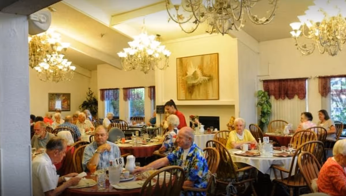 Elderly residents seated at round dining tables in a well-lit room with chandeliers.