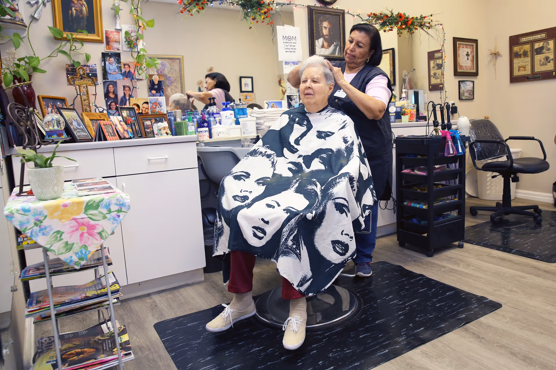 An elderly woman sits in a salon chair with a black and white cape featuring artistic faces, while a hairstylist stands behind her, cutting her hair. The salon is decorated with numerous framed photos, plants, and hair care products on the counter. A mirror reflects part of the scene, and the floor has black mats under the chairs.