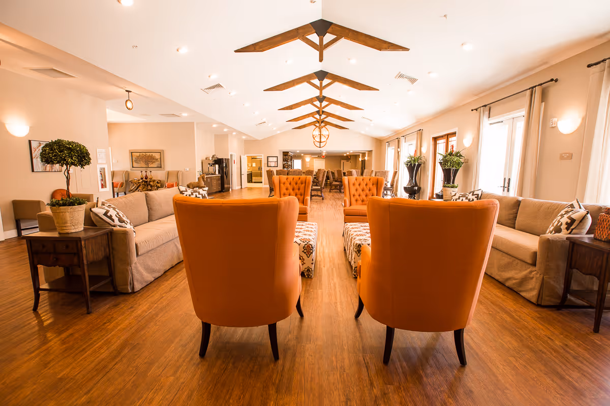 Spacious and well-lit senior living facility common area with wooden flooring, beige walls, and a high ceiling featuring wooden beams. The room is furnished with orange armchairs, beige sofas with patterned cushions, wooden side tables, and decorative plants. Large windows with curtains allow natural light to fill the space.