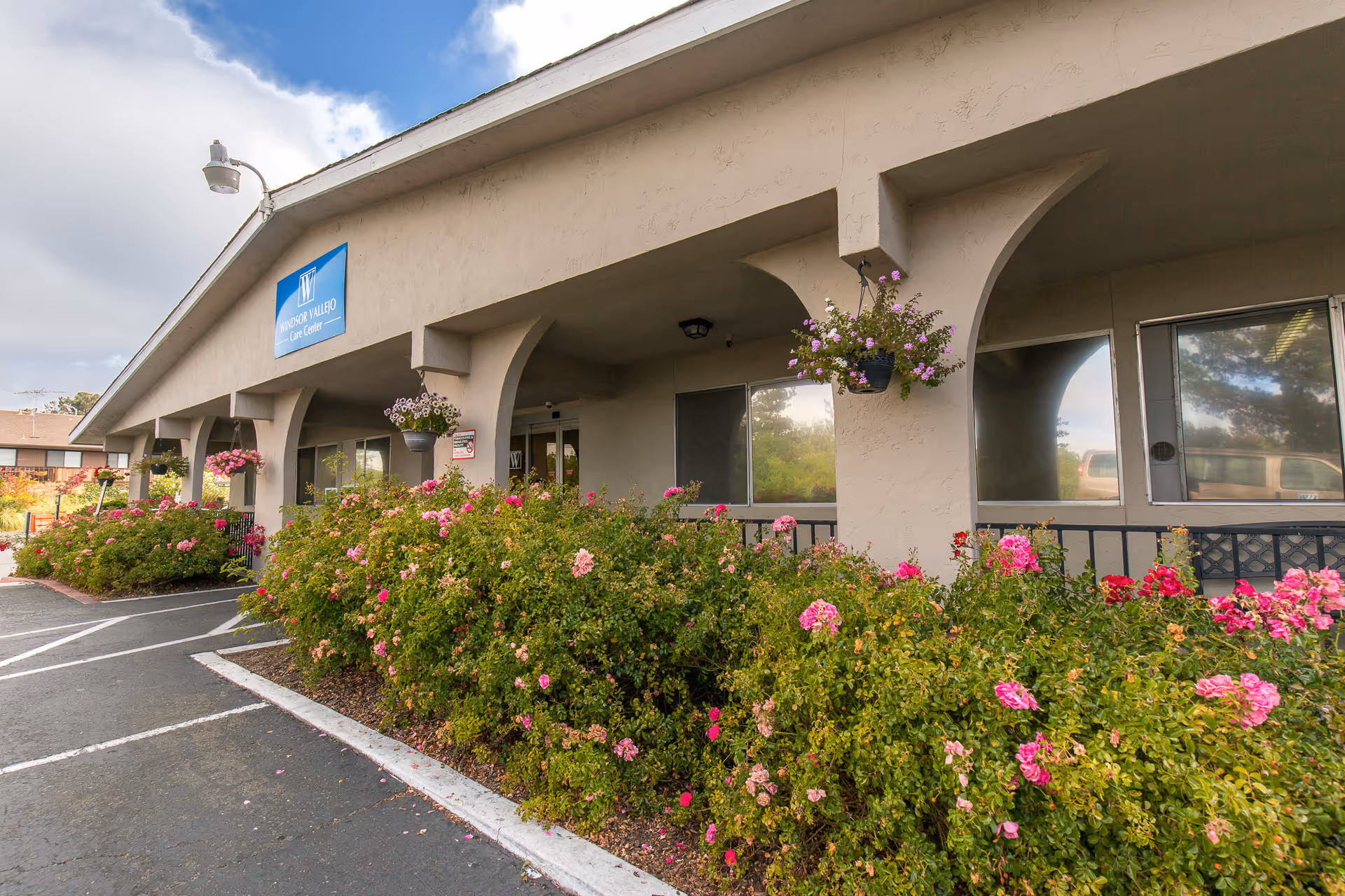 Exterior view of Windsor Vallejo Care Center building with beige walls, arched columns, hanging flower pots, and a garden bed filled with pink and red flowers along the sidewalk and parking area.