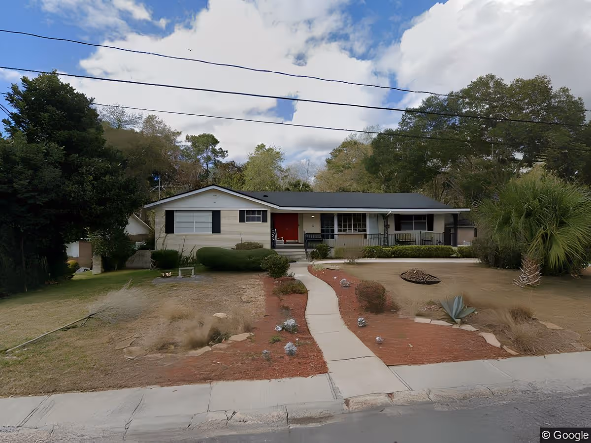 Single-story house with a front yard, walkway, and trees under a partly cloudy sky.