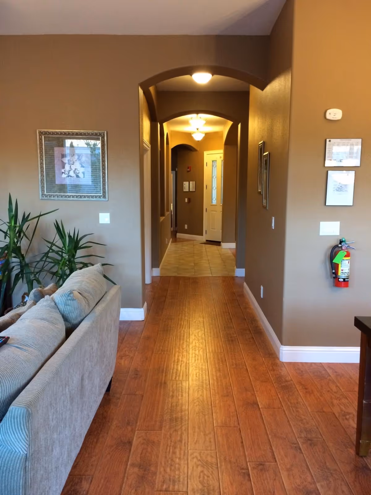 Interior view of a hallway in a senior living facility with wooden flooring, beige walls, and arched doorways. A beige couch with cushions is partially visible on the left side, along with some green plants. The hallway leads to a white door at the end, with framed pictures and a fire extinguisher mounted on the walls.