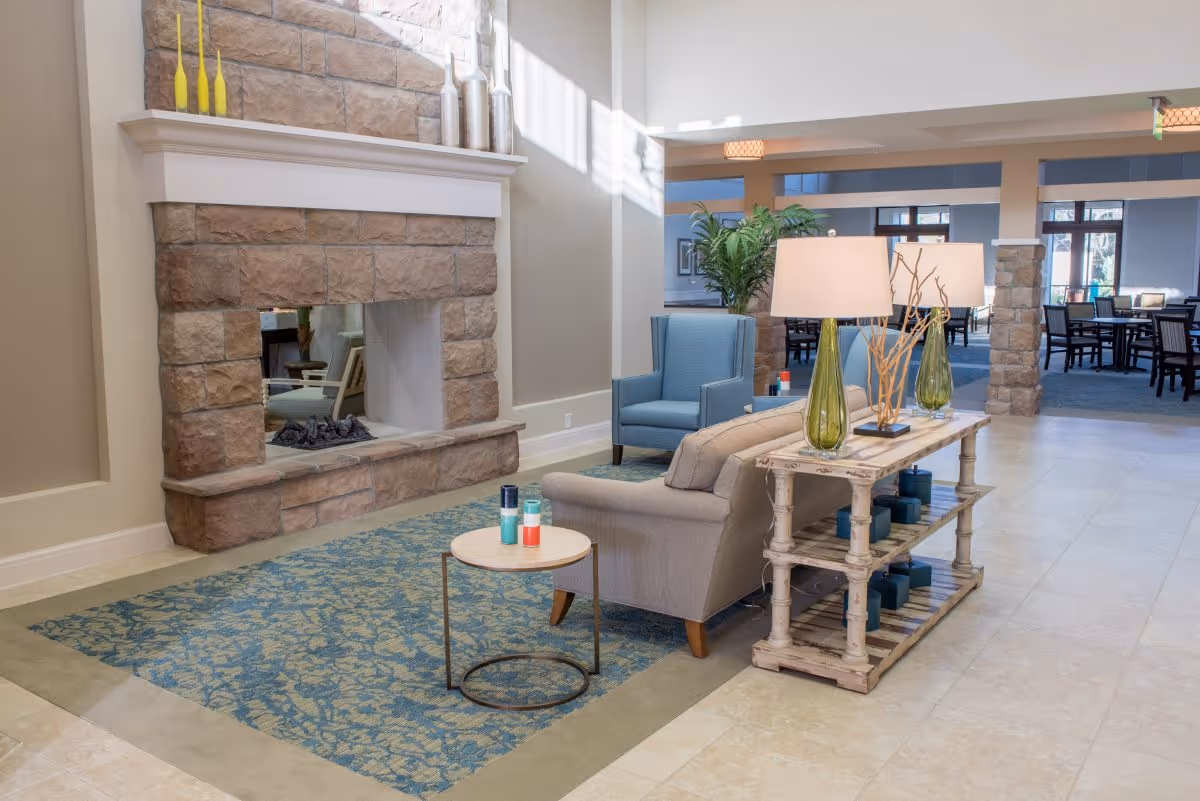 A cozy seating area in a senior living facility featuring a stone fireplace, a beige sofa, a blue armchair, a small round side table with decorative items, and a wooden console table with two green glass lamps and decorative branches. The background shows a dining area with tables and chairs, large windows, and stone pillars.