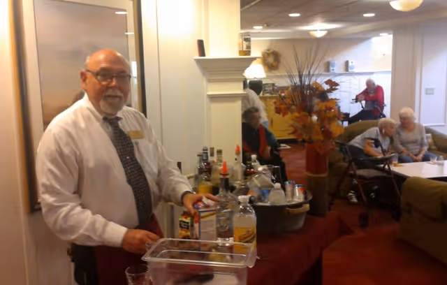 A staff member stands behind a drink station in a senior living lounge while elderly residents sit on couches nearby.