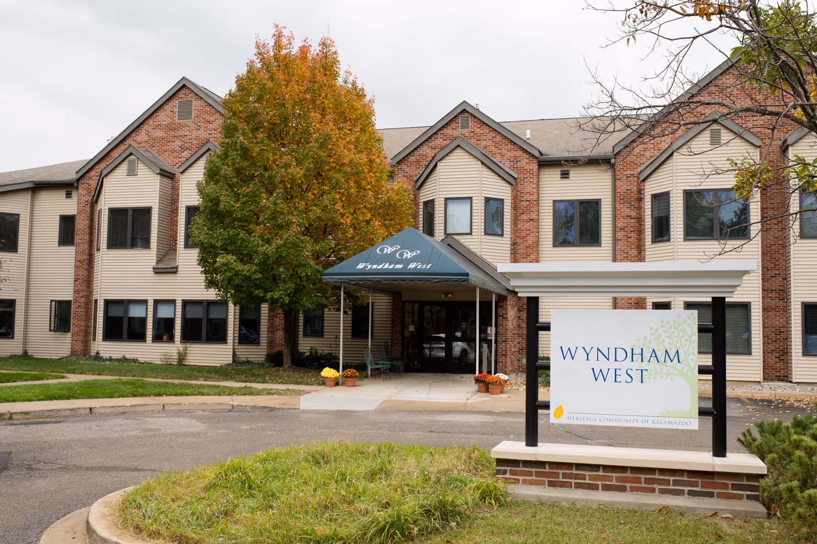 Exterior view of the Wyndham West building at Heritage Community of Kalamazoo, showing a two-story structure with beige siding and red brick accents, a green awning over the entrance, a tree with autumn foliage, and a sign in front displaying the facility name.