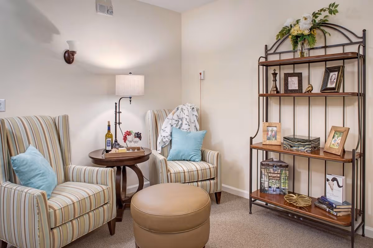A cozy sitting area with two striped armchairs each with a light blue pillow, a round wooden side table between them holding a bottle of wine, two wine glasses, and a small tray with grapes and cheese. A floor lamp is behind the table. To the right, there is a metal and wood shelving unit decorated with framed photos, books, a decorative box, and a flower arrangement on top. The walls are light beige and the floor is carpeted.