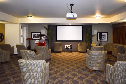 A cozy media room with multiple cushioned armchairs arranged facing a large white projection screen. The room has patterned carpet flooring, two tall plants flanking the screen, framed black and white photos on the walls, and a ceiling-mounted projector. There is also a red popcorn machine on the left side of the room.