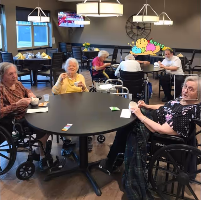 A group of elderly people sitting around tables in a dining area, some in wheelchairs, enjoying food and drinks. The room has modern lighting fixtures, a large wall clock, and a television mounted on the wall.