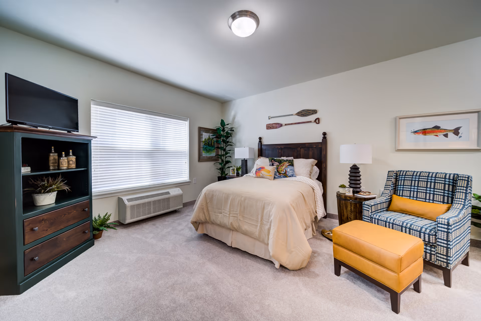 A cozy bedroom with a bed featuring beige bedding and decorative pillows, a wooden headboard with two paddles mounted above it, a patterned armchair with a yellow cushion, and a matching yellow ottoman. There is a wooden nightstand with a lamp and small plant, a green cabinet with a TV on top, and a large window with blinds. The room has light-colored walls and carpeted floor.