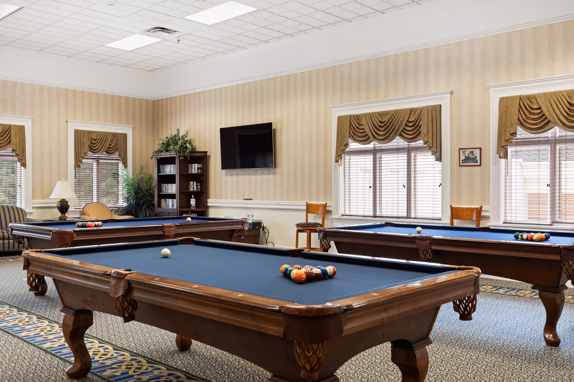 A spacious game room with three wooden pool tables with blue felt tops, arranged on a patterned carpet. The room has beige striped wallpaper, large windows with brown valances, a wall-mounted flat screen TV, a bookshelf with DVDs, and comfortable seating including a striped armchair and a wooden chair. There is also a table lamp and some greenery in the corner.