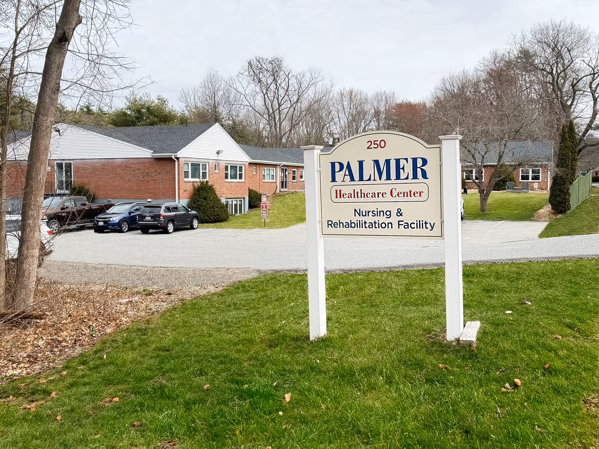 Outdoor view of Palmer Healthcare Center showing a white sign with blue and red text reading '250 PALMER Healthcare Center Nursing & Rehabilitation Facility' on a grassy area. In the background, there are brick buildings, parked cars, leafless trees, and a cloudy sky.