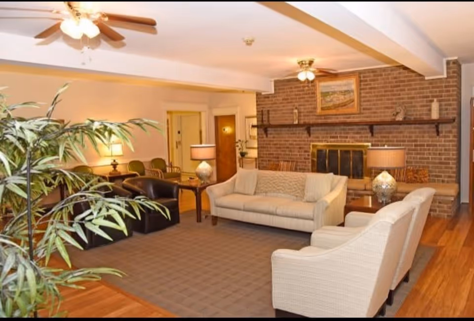 A cozy living room with a beige sofa, two matching armchairs, a dark brown leather chair, and a wooden coffee table. The room features a brick fireplace with a wooden mantle holding decorative items and a framed painting above it. There are two ceiling fans with lights, table lamps, and a large indoor plant in the foreground. The floor is wooden with a large area rug in the seating area.