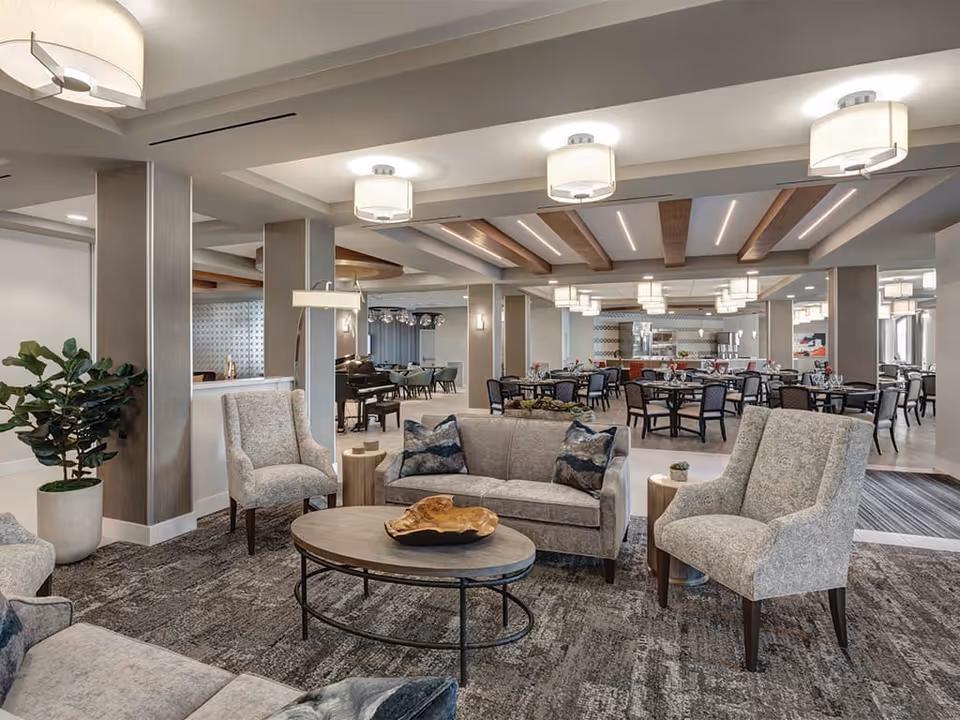 A spacious and well-lit common area in a senior living facility featuring comfortable seating with sofas and armchairs arranged around a round coffee table. The room has modern ceiling lights, decorative wooden ceiling beams, and a carpeted floor. In the background, there are dining tables and chairs set up for meals, along with a piano and large windows allowing natural light.
