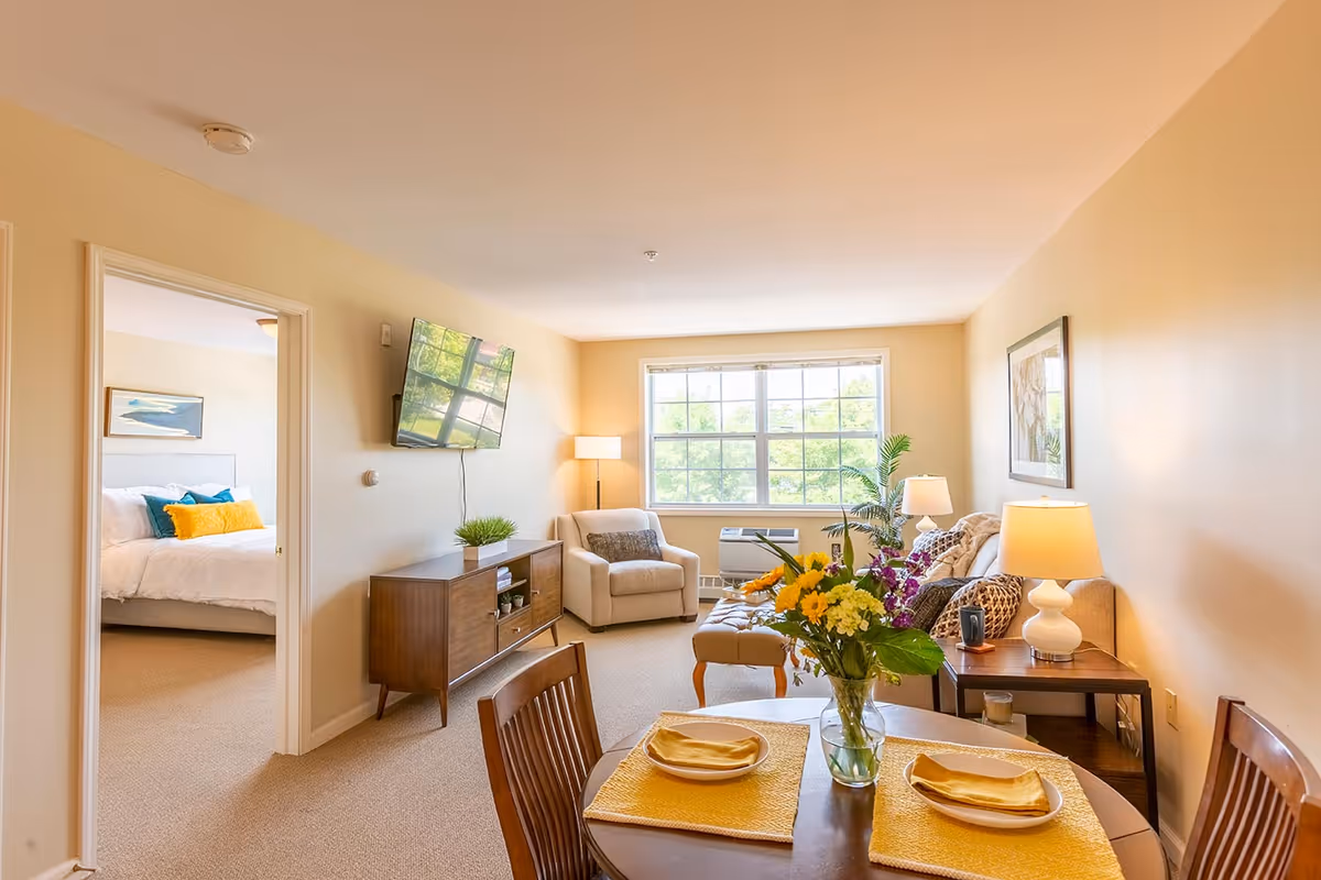 A bright and cozy living room with a large window letting in natural light. The room features a beige armchair, a wooden TV stand with a flat-screen TV mounted above it, a side table with a lamp, and a sofa with cushions. In the foreground, there is a round dining table set with yellow placemats, plates, and napkins, and a vase of colorful flowers. A doorway on the left leads to a bedroom with a neatly made bed and decorative pillows.