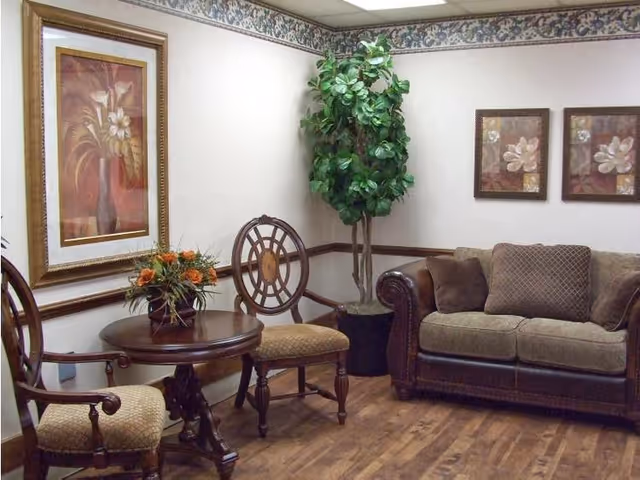 A cozy sitting area with a brown cushioned sofa, two wooden chairs with patterned cushions, and a round wooden table with a floral arrangement. The room has wooden flooring, a potted artificial tree in the corner, and three framed floral paintings on the walls with a floral wallpaper border near the ceiling.
