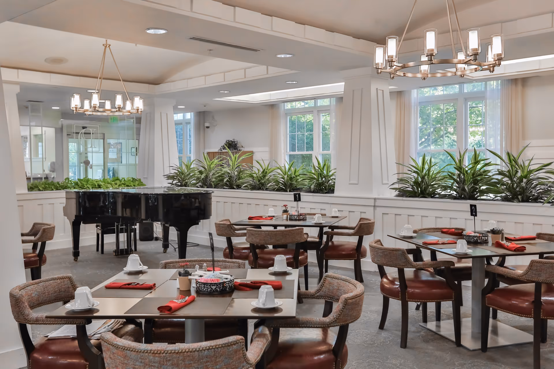 A bright and elegant dining room with multiple tables set with white cups and red napkins. The room features comfortable chairs, large windows letting in natural light, green plants along a white partition, and a black grand piano in the background under modern chandeliers.