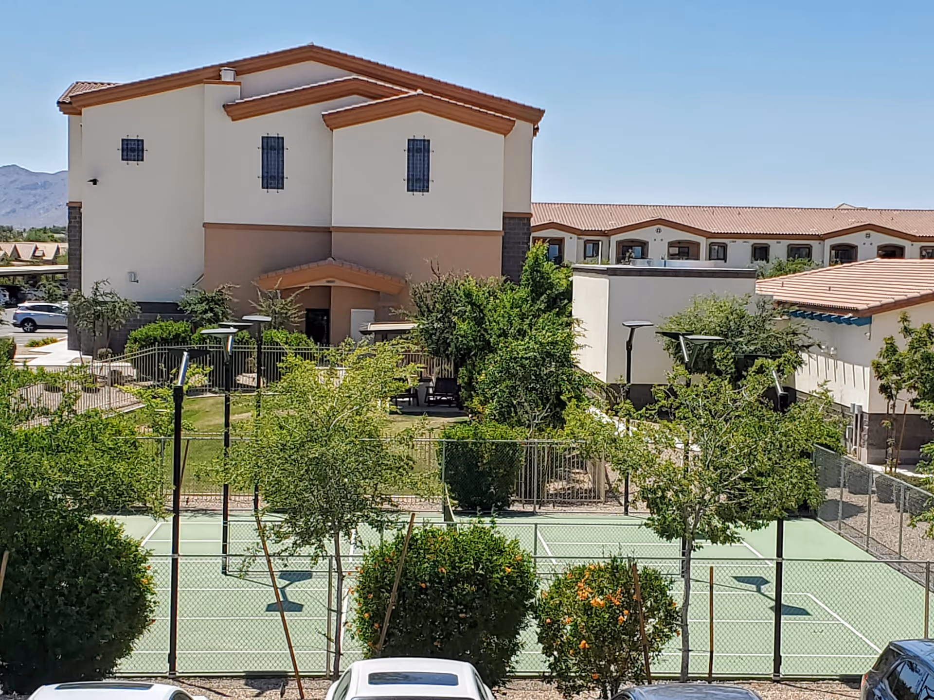 Exterior view of a multi-story senior living building with a fenced outdoor court, trees and parked cars in the foreground.