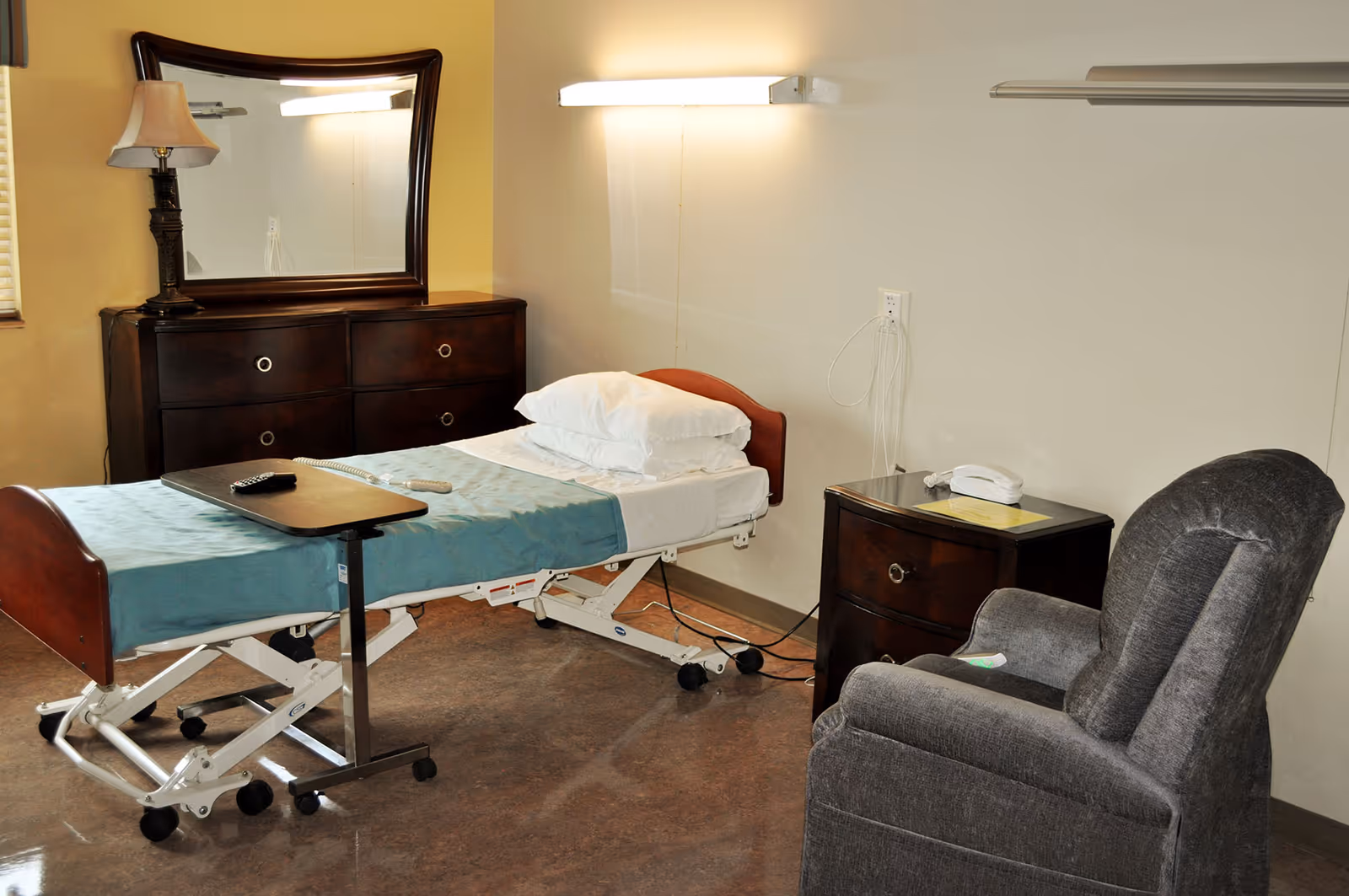 A room with a hospital-style adjustable bed covered with a teal blanket and white pillows. Next to the bed is a dark wooden nightstand with a white telephone on top. There is a dark wooden dresser with a large mirror and a lamp on it. A gray upholstered recliner chair is positioned near the bed. The room has beige walls and a polished floor.