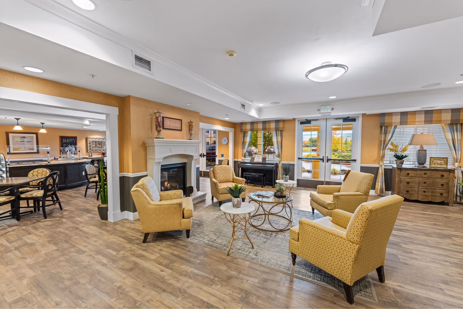 A bright and inviting senior living common area with four yellow armchairs arranged around two round coffee tables on a rug. There is a white fireplace with decorative items on the mantel, a piano near the window, and large windows with striped curtains letting in natural light. The room has wood flooring and leads into a dining area with tables and chairs.