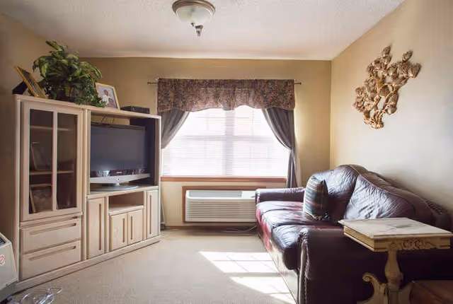 A cozy living room with a large window covered by curtains and a valance, a dark leather couch with a striped pillow, a wooden side table, and a light-colored entertainment center holding a flat-screen TV and some decorative items.