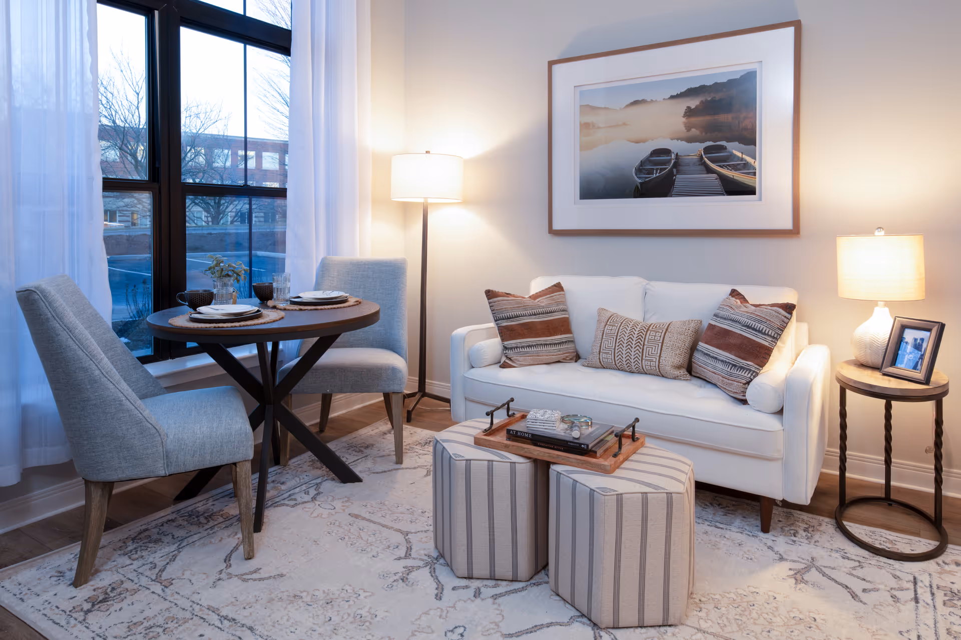Cozy living room with a white sofa, patterned pillows, framed artwork, a round dining table and chairs by a window, lamps, and striped ottomans on a rug.