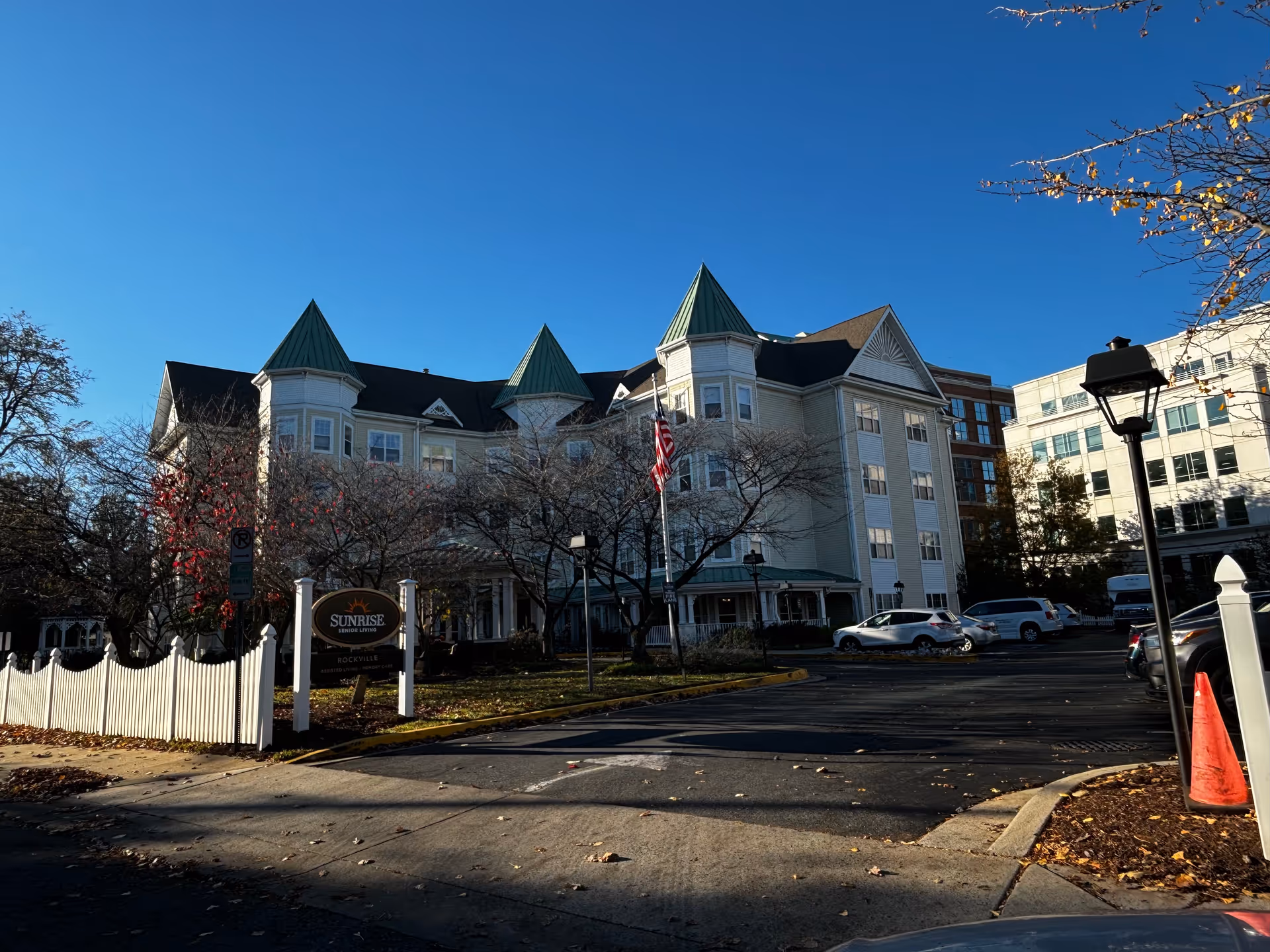 Sunrise of Rockville building exterior with green-roofed turrets, an American flag, white picket fence and a parking area.