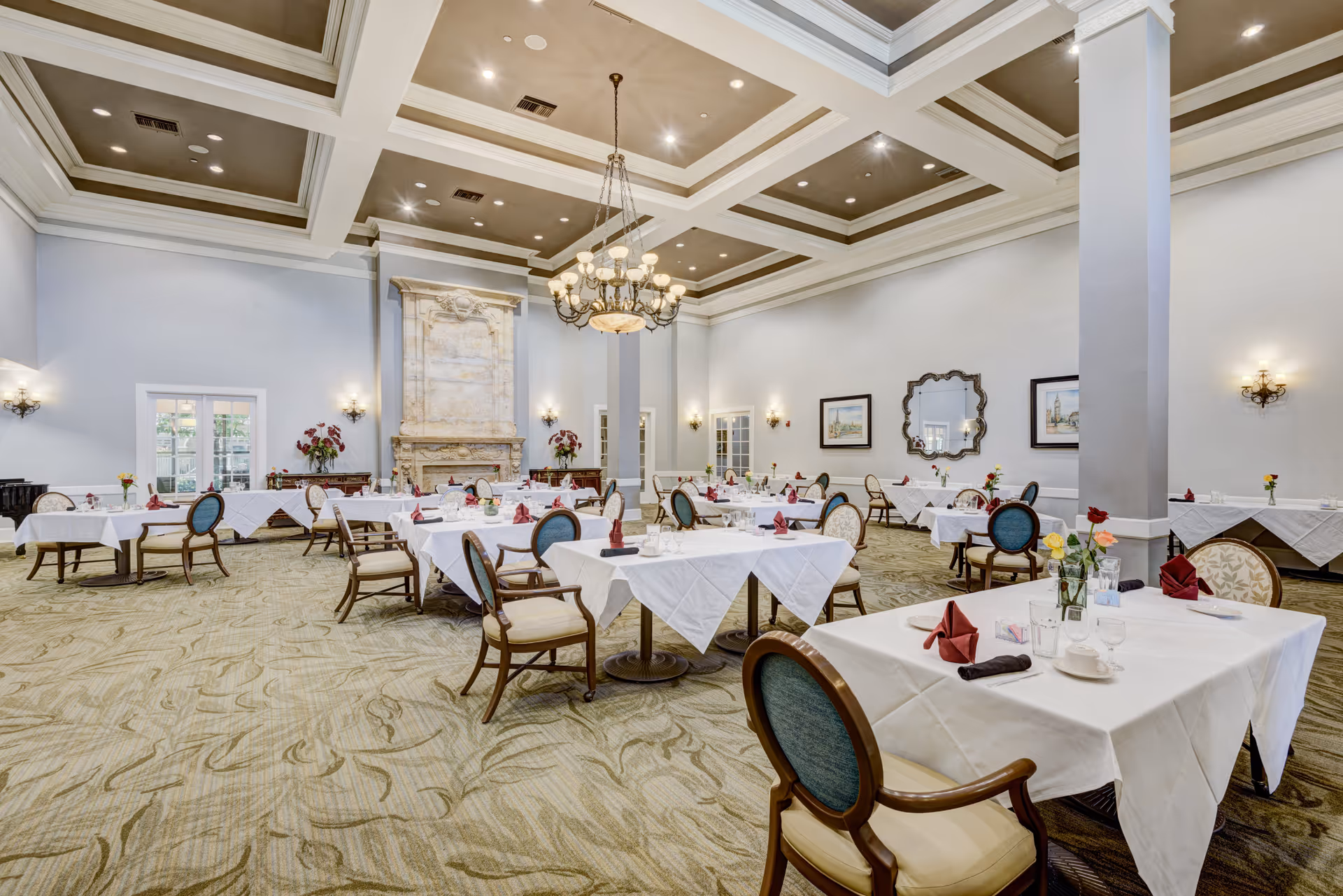 A spacious, elegant dining room with multiple tables covered in white tablecloths, each set with glasses, plates, and folded red napkins. The room features a high coffered ceiling with recessed lighting and a large chandelier. There is a decorative stone fireplace on one wall, framed artwork, a large ornate mirror, and floral arrangements on tables and sideboards. The carpet has a subtle patterned design, and the chairs have wooden frames with upholstered seats and backs.