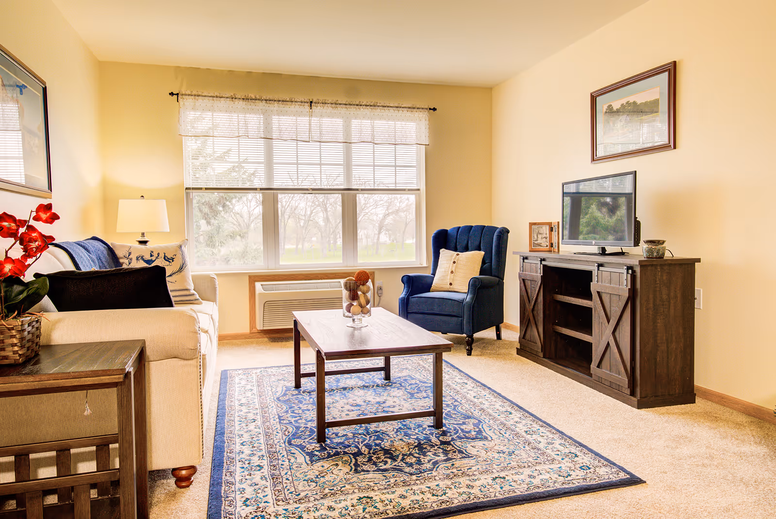 A cozy living room with a beige sofa adorned with pillows and a blue throw blanket, a dark wooden side table with a red flower arrangement, a blue upholstered armchair with a beige pillow, a wooden coffee table with decorative balls in a glass vase, a patterned blue and beige rug, a wooden TV stand with a flat-screen TV, and a large window with white blinds letting in natural light.