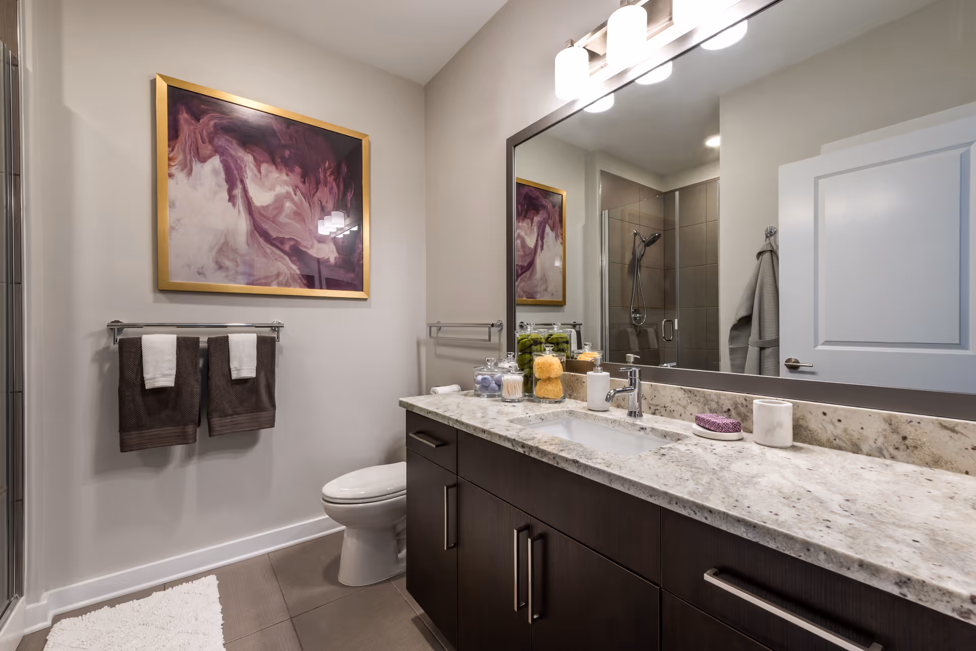 A modern bathroom with a granite countertop vanity featuring a sink, soap dispenser, and decorative jars. Above the vanity is a large mirror with three light fixtures. To the left, there is a toilet beneath a towel rack holding two brown towels with white accents. A framed abstract artwork with purple and white tones hangs on the wall. In the background, a glass-enclosed shower with a chrome showerhead is visible, along with a white door and a hanging towel.